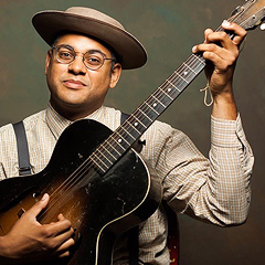 Dom Flemons, a thirty-something Black man holds a battered guitar, wearing an old-fashioned hat and shirt with suspenders, and round-rimmed spectacles.