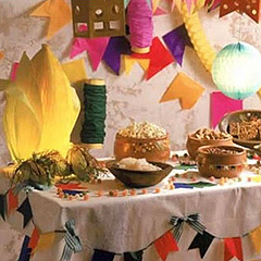 A table decorated for the Brazilian Feast of St John, with colorful banners and lighted paper lanterns.  The table holds an array of traditional foods in bowls and baskets.