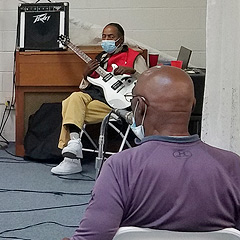A middle-aged Black man with an electric guitar is seated near an amplifier, and another middle-aged Black man is in the foreground, facing the guitarist and away from the camera.