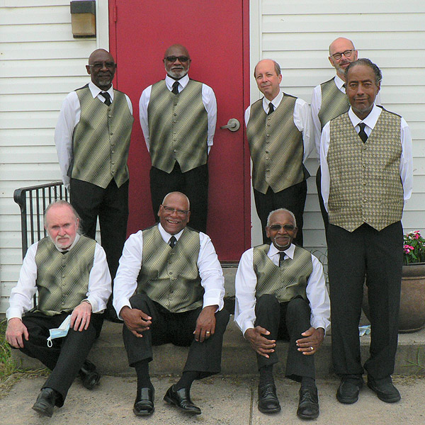 The eight members of the Spiritual Messengers pose for a group photo in their performance clothes of black slacks, white shirts, black ties and vests that are greenish-grey with a moiré pattern. They are all men, appearing to be in their 60s or older.  Five are Black; three are White. Most are smiling slightly, though Wayne Boulware (center front) is grinning. They are on the front stoop of their home-base church; five are standing and three are seated on the step.  The door behind them is cranberry red, and the siding is white.