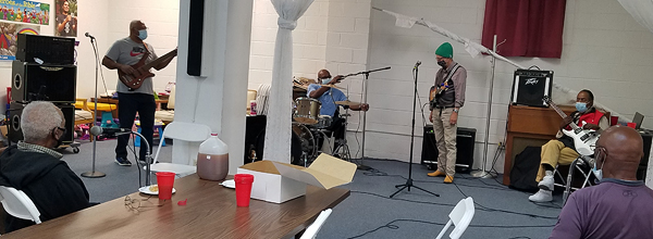 An informal rehearsal in the church’s fellowship hall. Rev. Jackson and Mac McFarland are singing from their seats at a table in the foreground; they are sitting with their backs to the camera.  On the far side of their large circle, four others are playing instruments: Wayne has his electric bass guitar, Anthony is sitting behind a drum kit; Tom has his mandolin, and Mark has his electric guitar. All the men are wearing masks and staying several feet apart from one another.