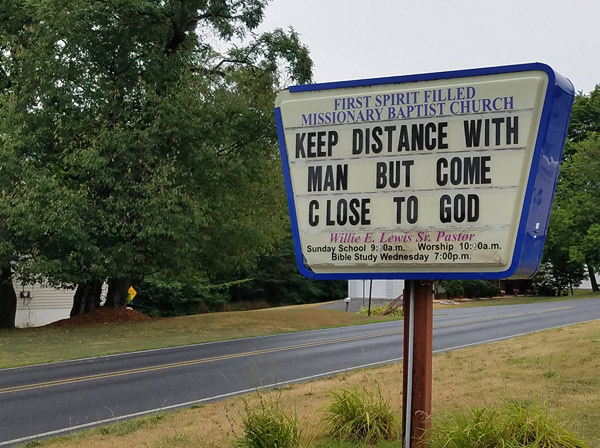 Trapezoidal sign by the road in front of the church: the name of the church appears at top, worship details at the bottom.  In the center, movable letters create this message: “Keep distance with Man but come close to God.”