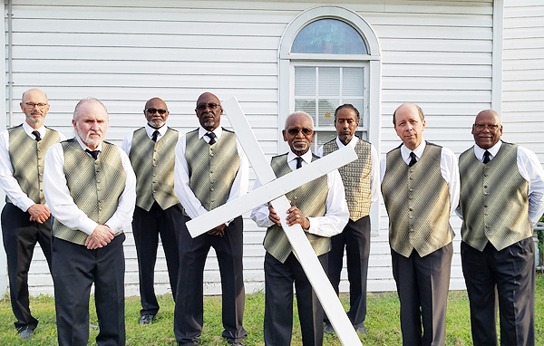 The Spiritual Messengers pose in front of their church.  Rev. Moses Jackson holds a large white cross in front of him, at an angle with one end on the ground.  The cross is made of 4-by-4 material and is taller than Rev. Jackson.