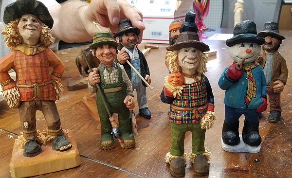An assortment of carved wooden caricature figurines, 6 to 8 inches high, with Jim's hand showing the scale. The characters are all male: a scarecrow, a fisherman, a halloween-themed scarecrow, a snowman's head on a human body, and a few others in the background.