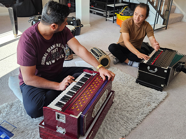 Two men sit on the floor, each with a harmonium, one finger on its keyboard and a hand on the bellows at the rear. The men are looking at one another. See first photo description.