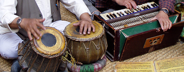 A pair of tabla drums and a harmonium are being played by two Nepalese men sitting on a sisal rug in Khatmandu. Only the men’s hands are visible.